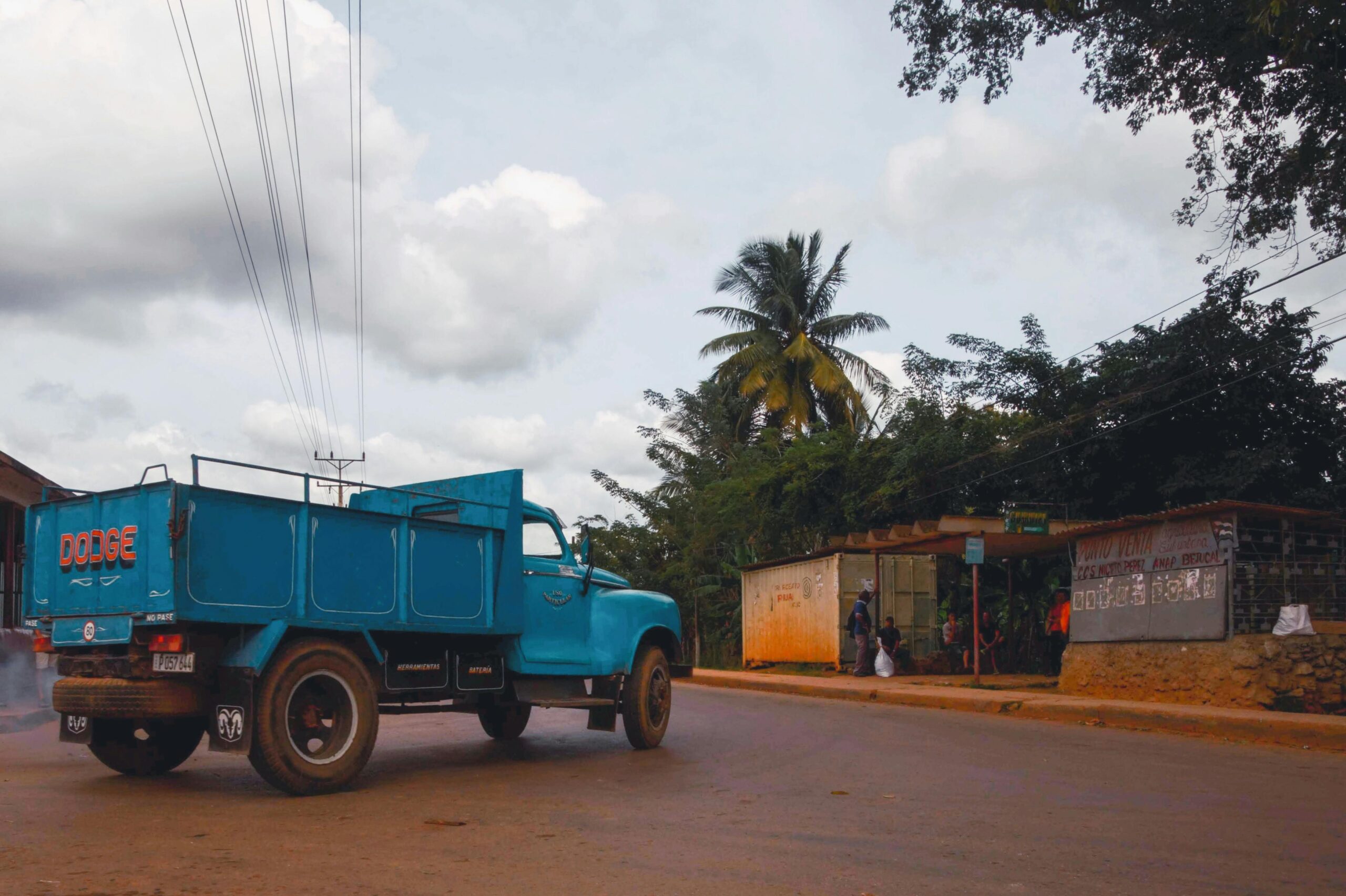 Old blue Dodge truck drives through a tropical urban street corner with palm trees and kiosks.